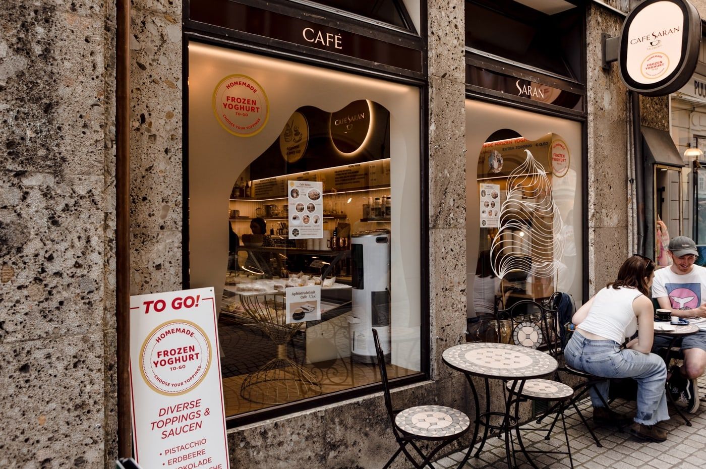 Das Café Saran von außen: Schaufenster mit dem Schriftzug Café, davor Mosaiktische, an denen Gäste in der Sonne sitzen
